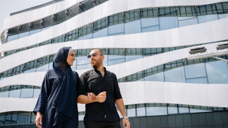 A couple stands happily together in front of a modern, wavy architectural building. The woman wears a dark blue hijab and matching abaya while the man is dressed casually in black with sunglasses. They are smiling at each other with joy as they embrace, surrounded by bright daylight and glass panels reflecting the sky behind them.