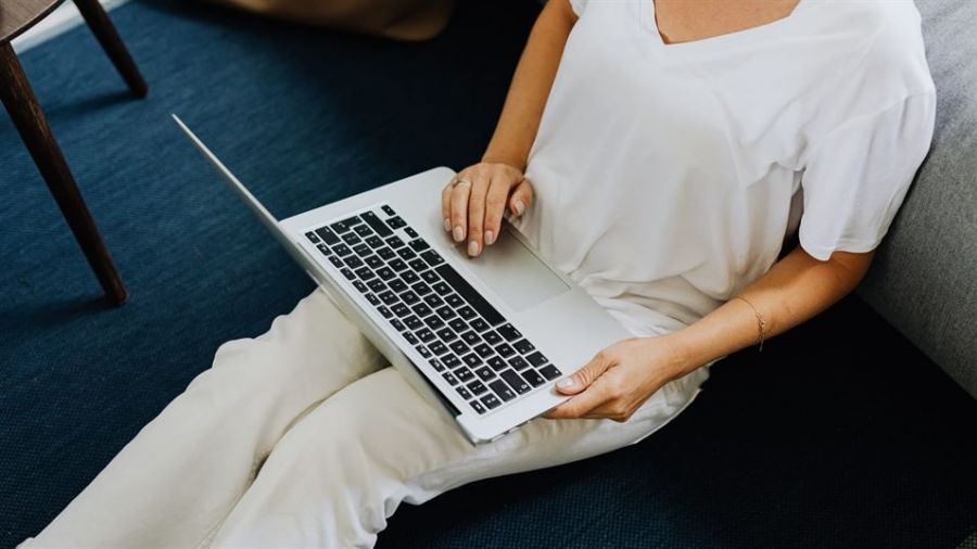 A person sits relaxed on a floor with their back against a couch while engaged with an open laptop on their lap. They wear a white T-shirt and light-colored pants, in a room with dark blue carpet that contrasts nicely with their outfit. The subtle lighting creates a calm atmosphere, making it look like they are working or enjoying leisure time at home under natural light.