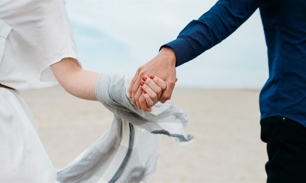 Uae Issues New Laws Why You Still Need To Get Married Dubai | New Marriage Laws In Uae - Why You Still Need To Get Married In Dubai | The New Marriage Laws In Uae Brings New Amendments To Its Family Laws. The New Laws Will Make It Easier For Non-National Unmarried People To Cohabitate. It'S Essential To Remember That These Laws Are Taking Effect Right Now Permitting The Unmarried Couple To Legally Share A Home. The image shows two people holding hands, symbolizing love and connection. They stand against a soft sandy backdrop with one dressed in white and the other in blue. The woman's red nails add brightness to this serene moment, showcasing joy and unity as they prepare for their wedding in Dubai.