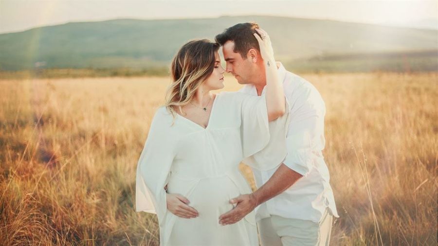 A couple stands lovingly in a golden field during sunset, holding each other with joy glowing on their faces. The woman wears a flowing white dress while she cradles her pregnancy bump, and the man gently rests his hand on it. They gaze into each other’s eyes, capturing a moment filled with love amidst a serene landscape of hills and blue skies. The intimate scene reflects warmth, connection, and anticipation for their shared future together.