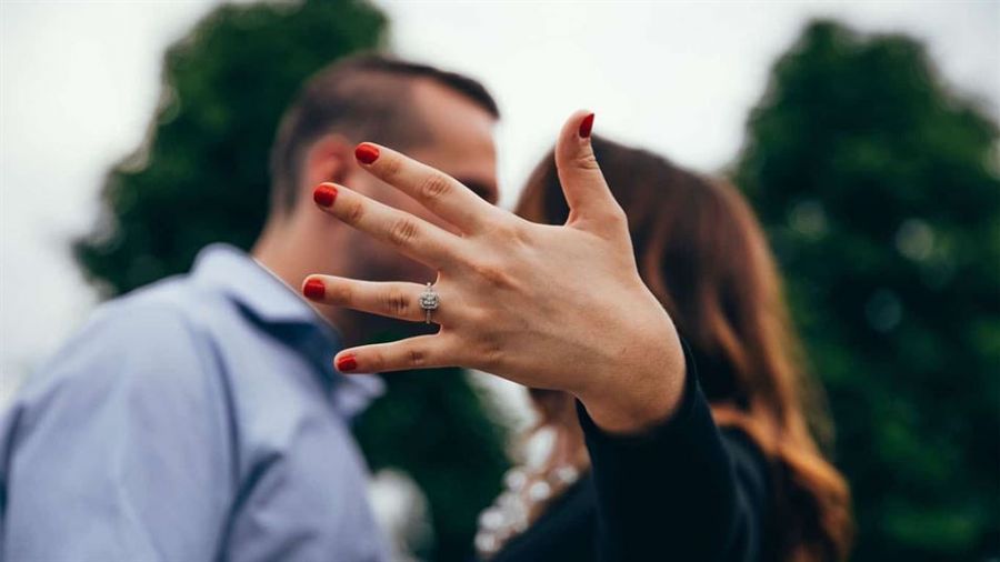 The image shows a happy couple outdoors with smiles as they celebrate an engagement moment. The woman has her hand raised displaying a sparkling engagement ring while wearing bright red nail polish. The background features soft green foliage creating a romantic atmosphere. Both appear joyful, capturing an intimate celebration together which signifies their commitment.