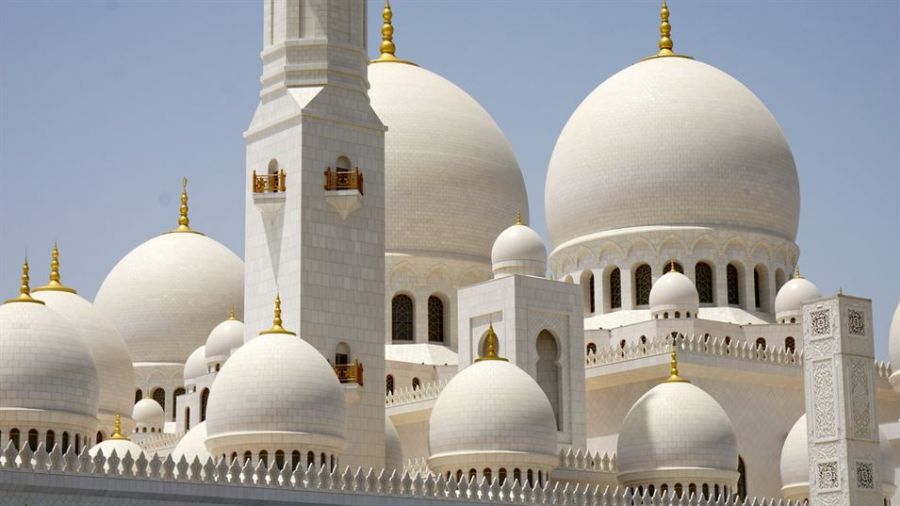 The image shows the Sheikh Zayed Grand Mosque in Abu Dhabi, featuring large white domes adorned with gold accents. The mosque's exquisite architecture creates a serene and majestic setting ideal for weddings. Clear blue skies highlight the beauty of this iconic structure, symbolizing tranquility and culture.