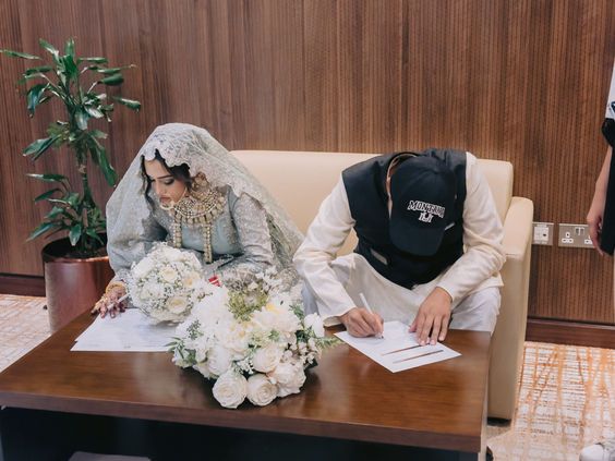 The image shows a couple happily sitting on a couch preparing for their wedding ceremony. The bride is wearing a beautiful white gown with delicate lace details while proudly holding a bouquet of white flowers. She has elegant jewelry and looks joyful as her groom, dressed in traditional attire, signs important marriage documents at their table filled with more floral arrangements. There's also a green potted plant that adds charm to the indoor space surrounding them.