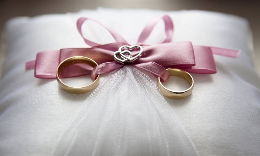 Get Married To Sponsor Your Partner In The Uae | Marriage For Sponsorship Recognized In Uae | There Is No Doubt That The Uae Has To Offer Things That Pretty Much Everyone Can Share. It'S One Of The Most Favored Places On Earth Where People Come To Live From All Over The World. But It Comes With Its Own Perks And Downsides That Everyone Has To Deal With. But Marriage Is One Of Those Things That Binds Two People Together To Help Each Other. Two gold wedding rings resting on a white satin pillow adorned with delicate tulle and topped with a pink ribbon bow. The bow features interlocking silver hearts at the center, symbolizing love and commitment. Soft textures create an elegant look, showcasing the romance of weddings.