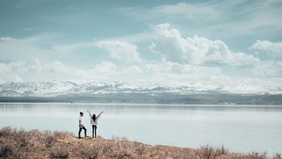 A beautiful image shows a serene view of two people by calm water. They are surrounded by fluffy clouds and mountains. One person raises their arms joyfully while the other admires the scenery. This captures a peaceful moment shared in nature, reflecting happiness and love in an enchanting setting.