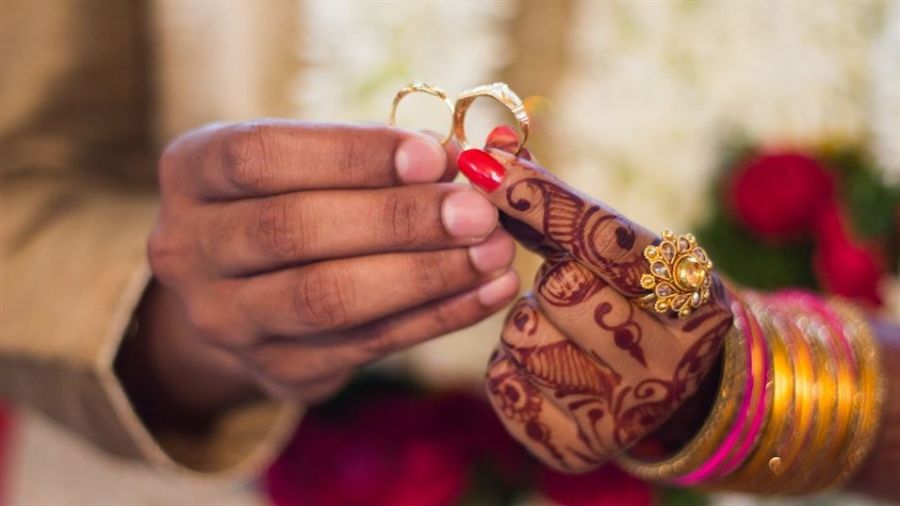The image displays two hands holding gold rings against a blurred floral background. One hand shows colorful bangles and intricate henna designs while presenting a ring delicately between the thumb and forefinger. This imagery symbolizes love and commitment within a wedding context.