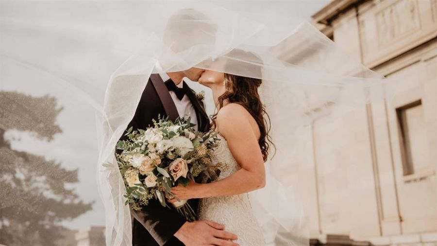 A couple shares a romantic moment under a delicate bridal veil during their outdoor wedding. The groom is wearing a black suit while the bride looks stunning in her white lace gown holding a bouquet of soft-hued flowers. The serene background features trees and elegant architectural elements, creating an inviting atmosphere for their special day.