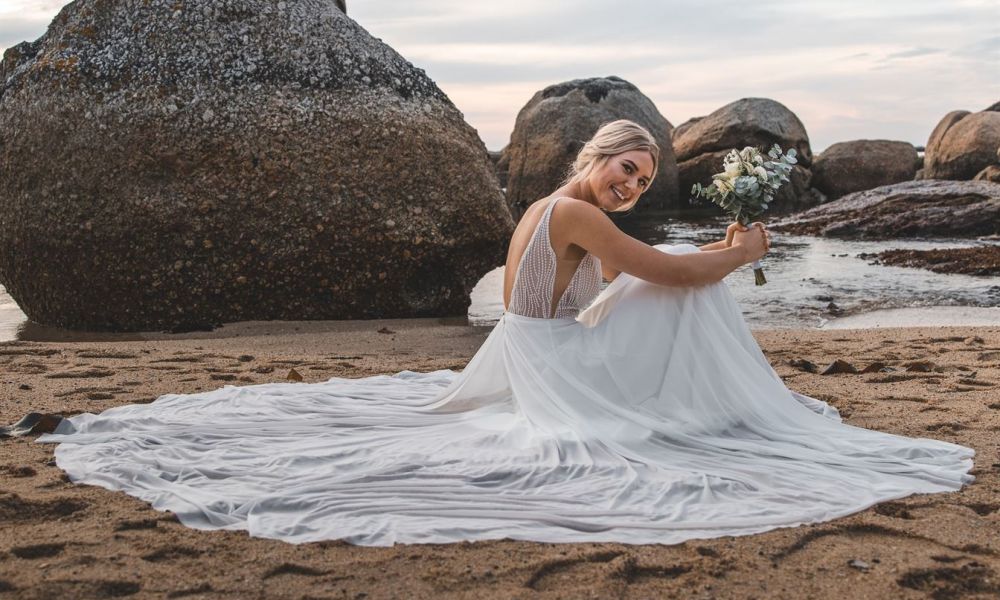 Can I Get Married In Georgia When You Live In Gcc Countries | Can I Get Married In Georgia When You Live In Gcc Countries | The Simple Answer Is 'Yes'. When A Couple Decides To Marry, The Ongoing Consideration Of What To Prioritize Might Lead To Discussions About What Is Best. Every Wedding Conversation Has Different Layers Just Like The Cake On The Wedding Day. The Best Version Of Everyone Will Always Be A Little Different. A bride sits gracefully on the beach wearing an elegant white dress and holding a bouquet of flowers. The ocean waves crash gently nearby while large rocks frame her serene moment. The cloudy sky adds to the tranquil beauty around her as she gazes peacefully into the horizon full of dreams.