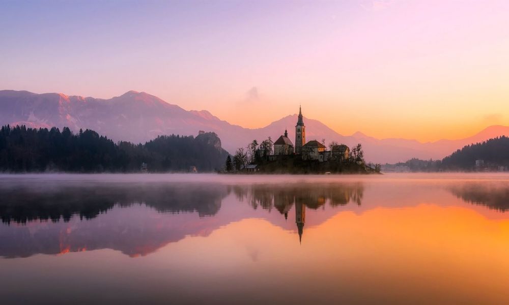 How To Get Married In Dubai Church | Everything You Need To Know About Planning Your Dream Wedding In Dubai Church | If You’re Looking To Plan Your Wedding In Dubai Church, Then You’re At The Right Place. The image shows a beautiful island at sunrise featuring a picturesque church surrounded by calm waters. Mist rises from the water creating an enchanting atmosphere. Silhouetted trees frame the scene while majestic mountains glow softly in pink and orange hues beyond them. The landscape conveys peace and natural beauty perfect for weddings or romantic escapes.