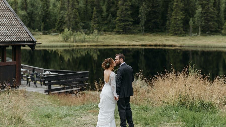 A happy couple stands by a serene lake, dressed in beautiful wedding attire. The bride is in a lovely white gown while the groom wears a sleek suit. They seem to be enjoying each other’s company surrounded by lush greenery and trees, creating a romantic atmosphere that captures the joy of love and celebration during their wedding ceremony.