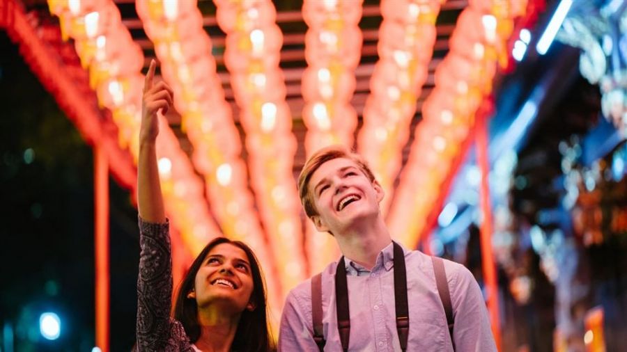 A happy couple stands together under bright red lanterns glowing softly around them. The woman joyfully points upward while the man smiles broadly at her side. They appear relaxed and casual as they share a delightful moment together in a festive atmosphere. The surrounding lights create a warm ambiance that highlights their happiness and love for each other amidst the vibrant scene.