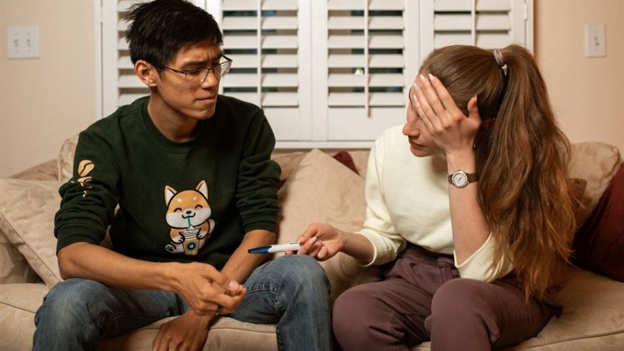 A couple sitting on a beige couch looks concerned as they discuss something important. The man wears a green sweatshirt and glasses while gesturing with his hand. The woman is in a white sweater, holding her forehead with one hand and showing him a pregnancy test with the other hand. They appear intimate and emotional, surrounded by soft lighting from window shutters behind them.