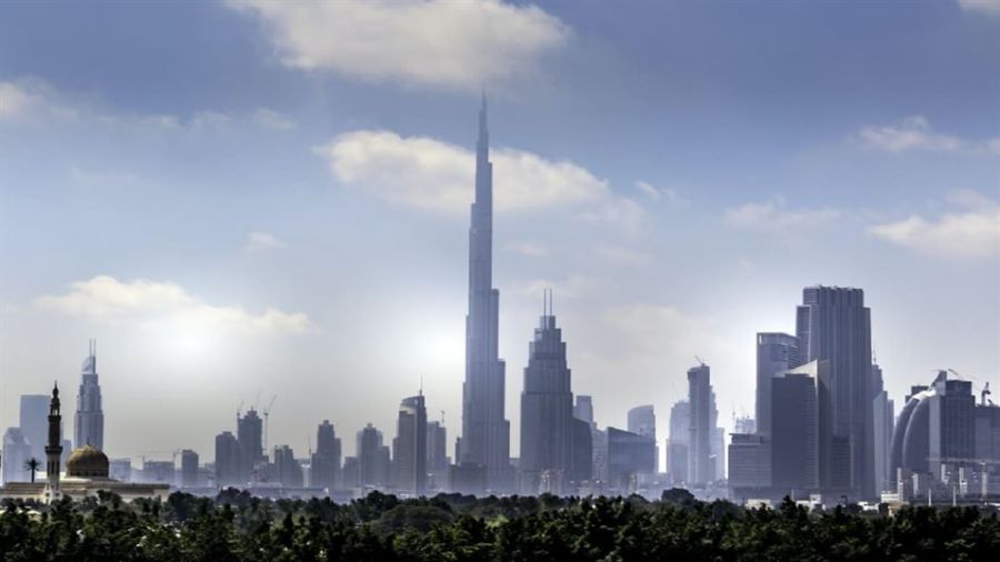 The image shows a beautiful view of Dubai's skyline featuring the Burj Khalifa surrounded by modern skyscrapers. The foreground has lush greenery which contrasts with the urban structures in the background. The sky is partly cloudy creating soft shadows on the buildings while highlighting their futuristic designs. This panorama captures Dubai’s dynamic essence as a global hub blending nature with impressive architecture offering a glimpse into its vibrant lifestyle.