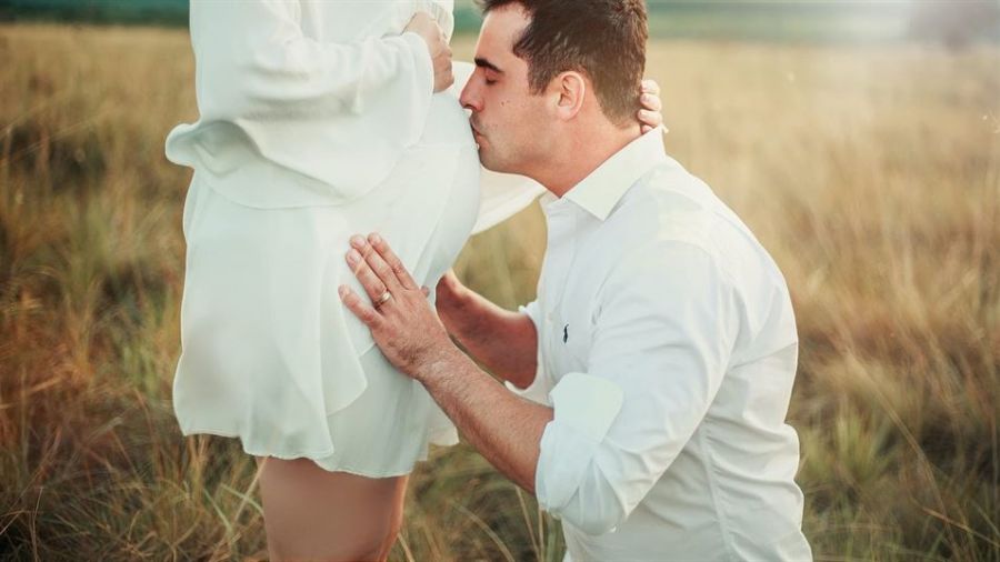 An image of a couple outdoors shows a man tenderly kissing the belly of a pregnant woman. They wear elegant white outfits amidst tall grass in soft sunlight, radiating love and serenity.