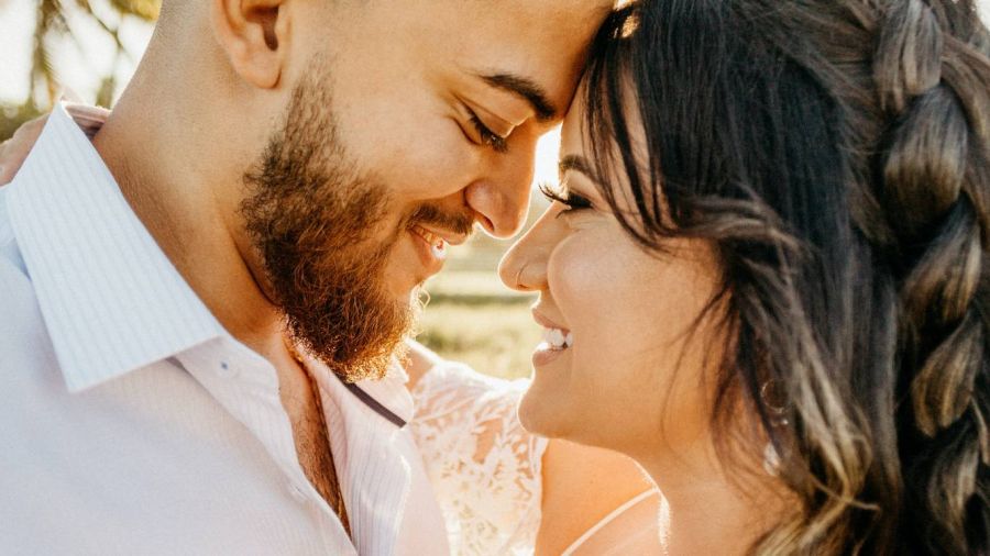 A happy couple leans together with their foreheads touching in an outdoor setting filled with soft natural light. The man has a beard and wears a light shirt while the woman in white lace shows her braided hair cascading down her back. Their smiles radiate warmth and love against a softly blurred background, encapsulating the joy of sharing their special moment together on their wedding day.