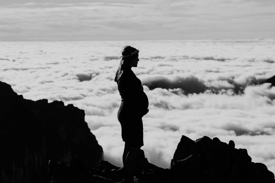 A silhouette of a pregnant woman in a dress stands on a rocky cliff, looking out over clouds. The scene captures her profile against a vast sky filled with fluffy white clouds. The contrast of darkness from the rocks and brightness from the clouds creates an emotional tone, portraying tranquility and reflection as she enjoys nature's beauty around her while contemplating life ahead.
