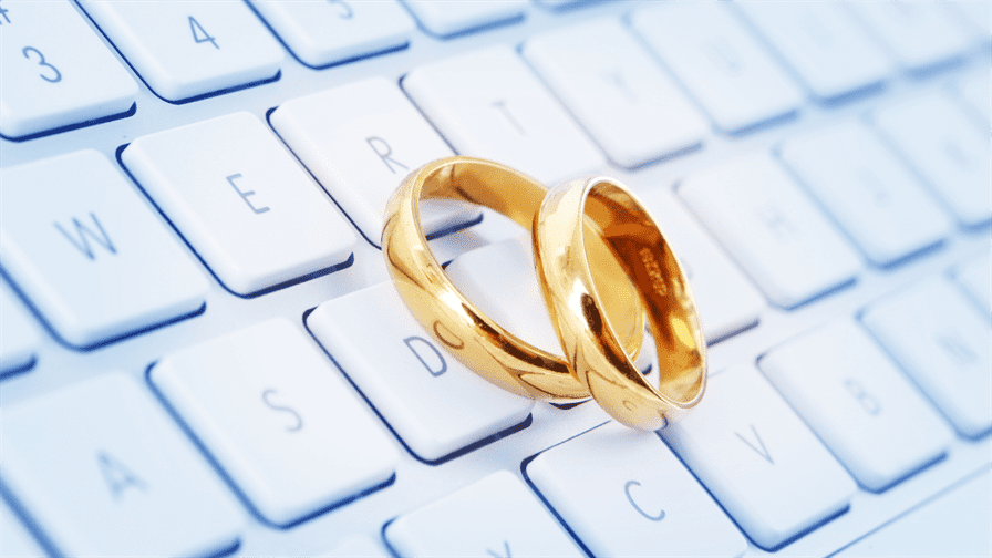 The image features two shiny gold wedding rings resting on a white computer keyboard, suggesting a blend of tradition and technology. The keys visible include letters such as W, E, D, and S which highlight themes of weddings. The bright background enhances the elegance of the rings while showcasing the digital connection to modern marriage processes.