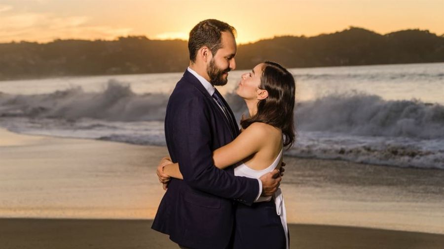 A happy couple embraces on a beautiful beach at sunset with gentle ocean waves and a golden sky behind them. The man wears a sleek dark suit while the woman shines in her elegant white dress. Their joyful smiles radiate love as they celebrate their special moment together in this romantic atmosphere filled with warmth and peace.