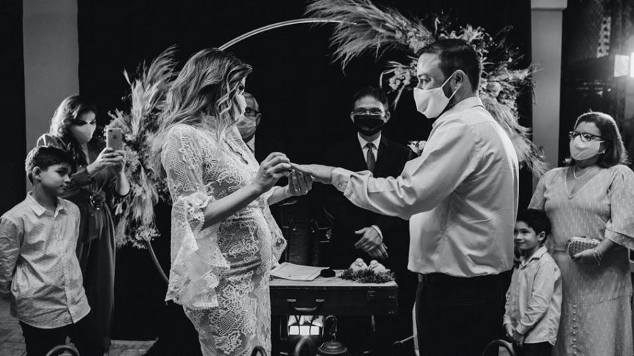 An intimate wedding ceremony scene showing a couple exchanging rings while wearing face masks. The bride looks lovely in her lace dress and the groom is dressed simply in a shirt and trousers. Surrounded by guests who also wear masks, they stand before an officiant under decorative floral arrangements. The ambiance highlights safety protocols during this special occasion while maintaining elegance and warmth with a charming arch behind them.