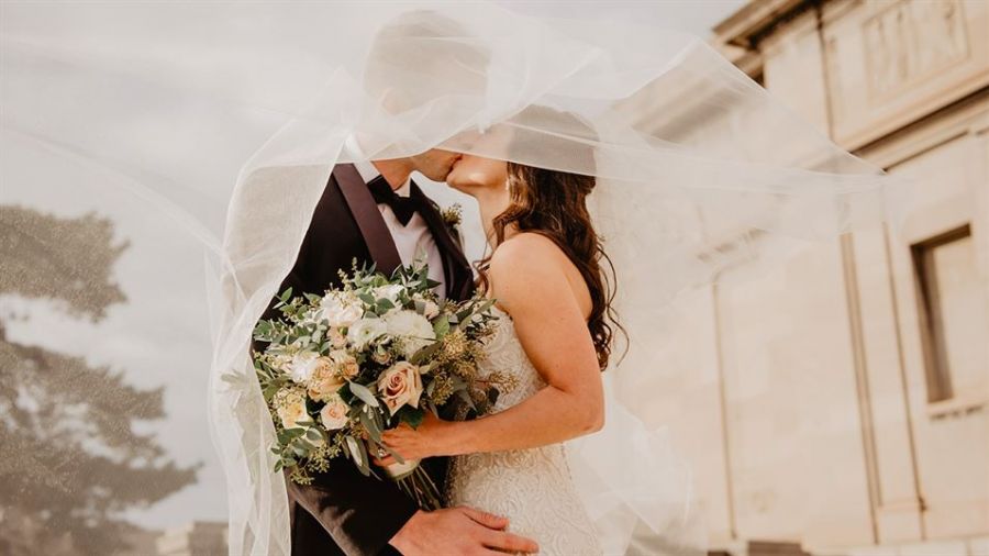 A romantic couple sharing a kiss on their wedding day outdoors. The bride wears a beautiful lace gown while holding pastel flowers. Her flowing veil glows softly in the sunlight, creating a dreamy effect around them. The groom looks dapper in his classic suit amidst an elegant architectural backdrop that indicates their wedding venue. This image captures the intimate joy of their celebration, making it perfect for couples dreaming of their own special moment in Dubai.