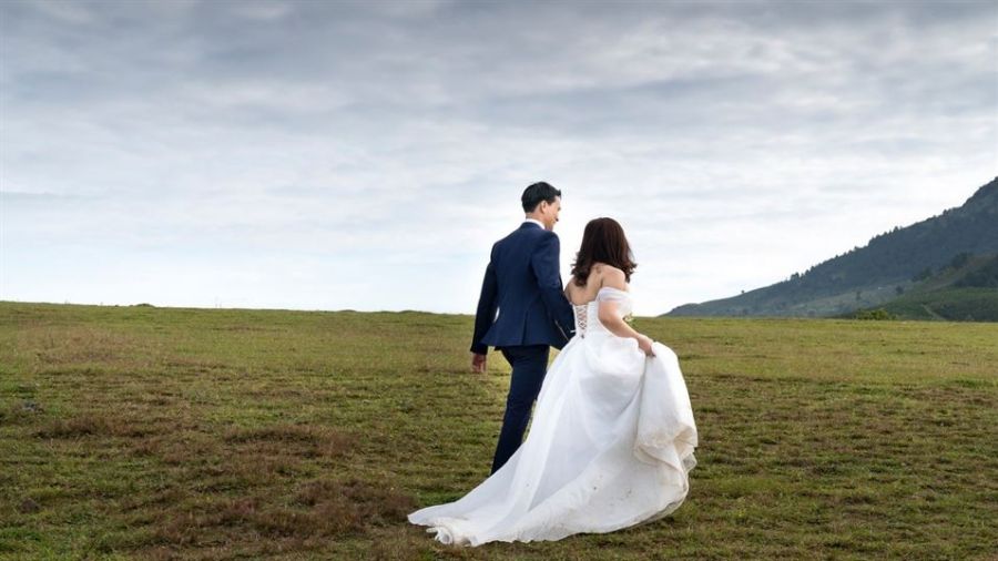 The image captures a couple joyfully walking hand in hand on a grassy landscape. The bride is adorned in a white gown with a long flowing train while the groom wears a dark blue suit. Their surroundings feature green rolling hills under an idyllic cloudy sky, creating a romantic outdoor atmosphere that symbolizes everlasting love.