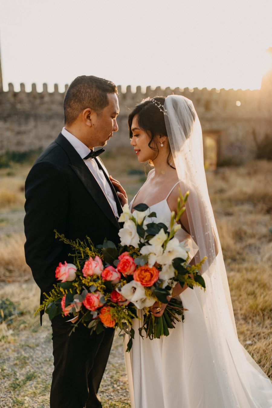 The image features a joyful bride holding a colorful bouquet of flowers beside her groom in a black tuxedo. They are surrounded by warm golden lighting that enhances the romantic vibe of their outdoor wedding setting. The couple looks deeply into each other's eyes showcasing love and happiness. Historical stone walls can be seen in the background creating a fairy-tale ambiance capturing the spirit of their special day.