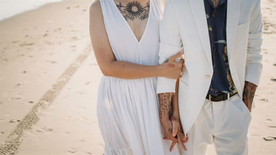 A happy couple walks hand-in-hand on a sandy beach showcasing a romantic moment. One is dressed in an elegant sleeveless white dress while the other wears a stylish white suit with a dark shirt. Both partners display unique tattoos adding personal flair as they enjoy their special day together near the water's edge with tire tracks visible in the sand suggesting recent arrival or fun times ahead.