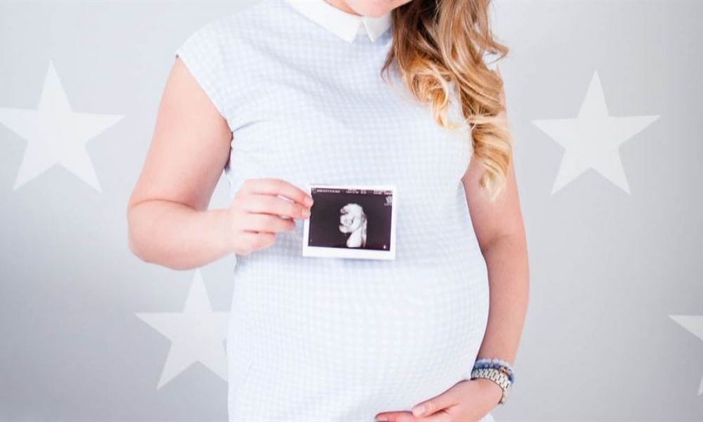 If You Are In The Uae And Are Pregnant But Not Married New Pregnancy Laws Apply To You | If You Are Pregnant But Not Married And Live In The Uae, New Pregnancy Laws Apply To You. | The Uae Is Constantly Making Positive Reforms When It Comes To Personal Laws. One Of The Biggest Reasons For These Changes Is To Attract Foreign Expats Who Often Look At The Middle East As The Hub For Investment And Business. The image features a pregnant woman wearing a light blue dress holding an ultrasound photo in front of her belly. She has loose wavy hair, adorned with several bracelets on her wrist. The background displays large white stars over a gray backdrop, enhancing the theme of motherhood and anticipation for new beginnings. This picture captures emotions of excitement and family warmth as she dreams about the upcoming arrival of her child.