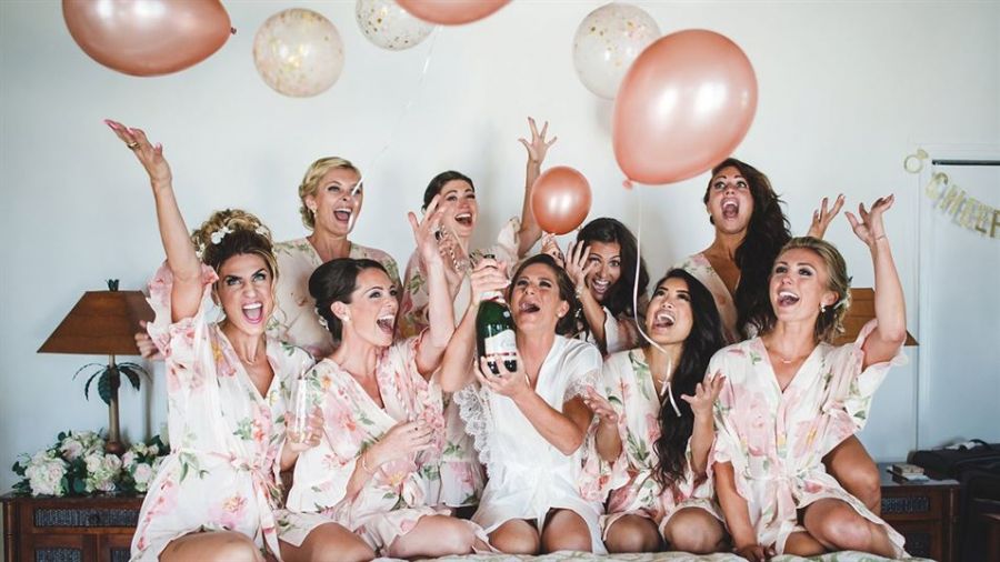 A joyful group of women dressed in matching floral robes celebrate together indoors with raised hands and big smiles. One woman holds a champagne bottle while pink and cream balloons float above them adding festivity. The backdrop features a bride-to-be banner enhancing the celebratory atmosphere typical of bridal showers or pre-wedding events. The overall scene radiates excitement, happiness, and camaraderie among friends preparing for a wedding celebration.