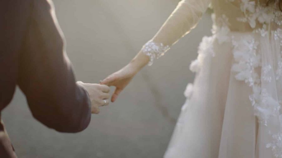 A close-up image of a couple holding hands, with one partner in a dark suit and the other wearing an elegant white wedding dress decorated with floral lace details. The setting is softly lit with warm sunlight, giving a romantic feel, while the background is blurred to focus on their connection. This image symbolizes love and partnership during wedding ceremonies in Dubai or Abu Dhabi.