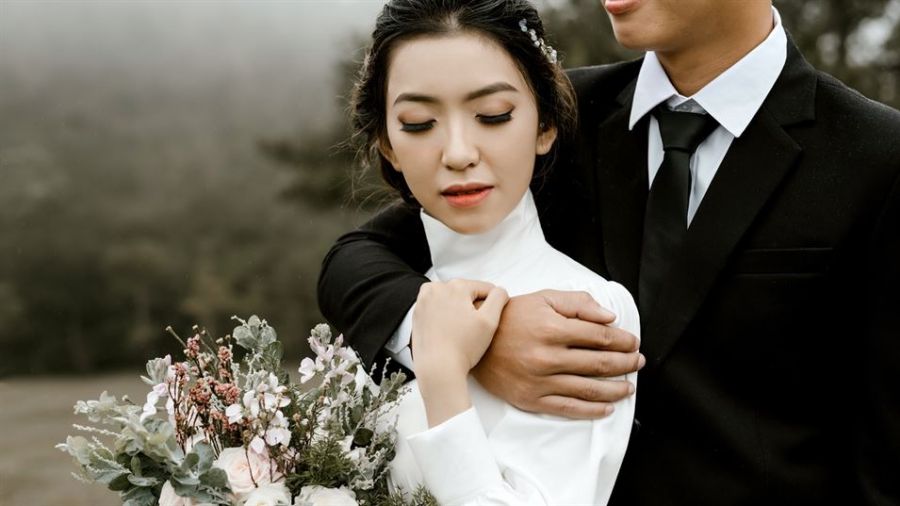 A happy couple dressed in a white wedding dress and a black suit embrace each other tenderly outdoors, surrounded by soft greenery. The woman smiles peacefully while holding a bouquet of pink and white flowers. Their romantic moment is captured beautifully with a dreamy blurred background, creating an atmosphere filled with love and joy perfect for weddings in Dubai.