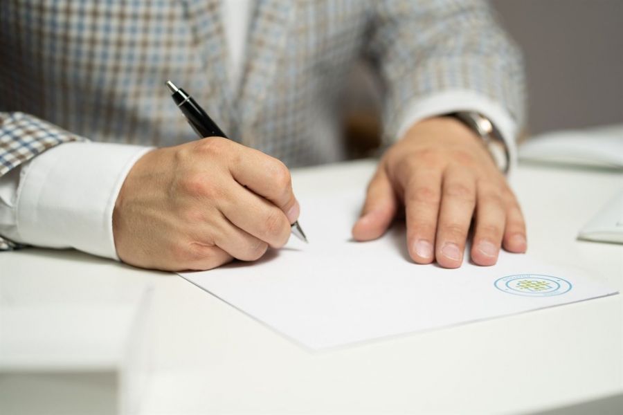 An individual sits at a white table and writes with a black pen on a document that has an official emblem. They wear a stylish checkered blue and white blazer over a light shirt. The scene conveys a formal atmosphere focused on completing marriage-related paperwork, emphasizing the significance of legal documentation in wedding preparations.