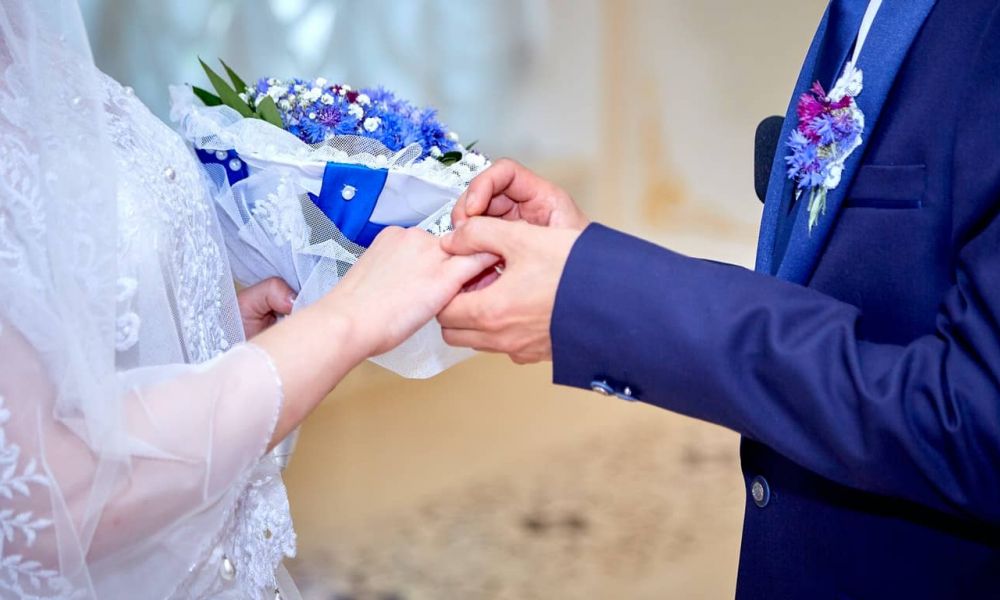 The Prerequisites For An Easy Wedding In Dubai | The Prerequisites For An Easy Wedding In Dubai | Recently, Dubai Has Seen An Increase In Expats Trooping Into The Emirate For Business And Relocation Purposes. Many Of These Expats Believe That The Emirate Holds A Lot Of Prospects. For This And Many Other Reasons, They Pitch Their Tents In Dubai. A lovely bride and groom share a tender moment as they exchange rings at their wedding ceremony. The bride wearing a delicate white dress holds a beautiful bouquet while the groom in a dark suit smiles lovingly. This image captures an intimate celebration filled with joy and warmth surrounded by elegant decor that enhances the romance of their special day in Dubai's magical atmosphere.