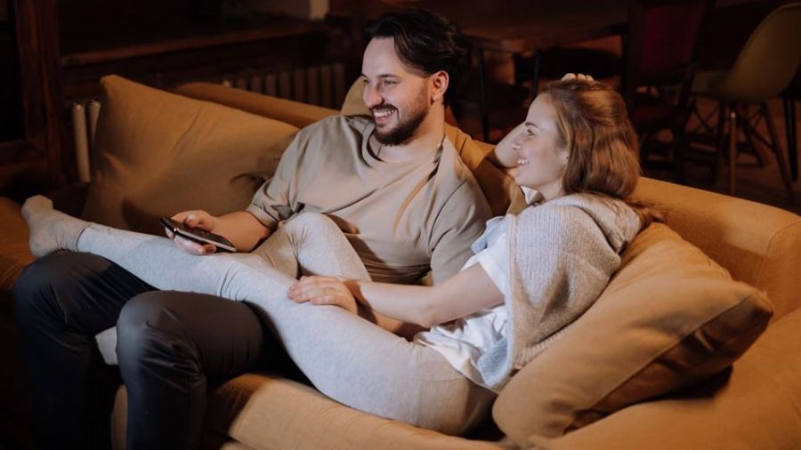 A happy couple is sitting on a light brown couch enjoying a cozy moment together. The man wears a light brown shirt while the woman is dressed in a comfy gray sweater. They appear relaxed and connected in their warmly lit living room, creating an inviting atmosphere perfect for couples considering marriage in Dubai.