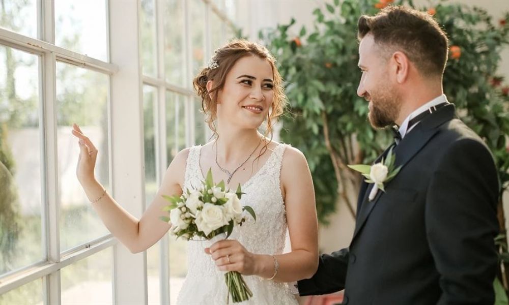 Is It Possible To Get Married On The Same Day In Georgia | Can Uae Expats Get Married On The Same Day In Georgia? | The Answer Is Yes ! At The Public Hall, You Can Lawfully Marry On The Same. An excited couple stands inside a bright room adorned with large windows and lush green plants. The bride wears a gorgeous white lace wedding dress, holding a bouquet of white flowers while smiling radiantly at her partner. He looks dapper in a dark suit with a bow tie. The light streaming through the windows creates an enchanting atmosphere for their special day.