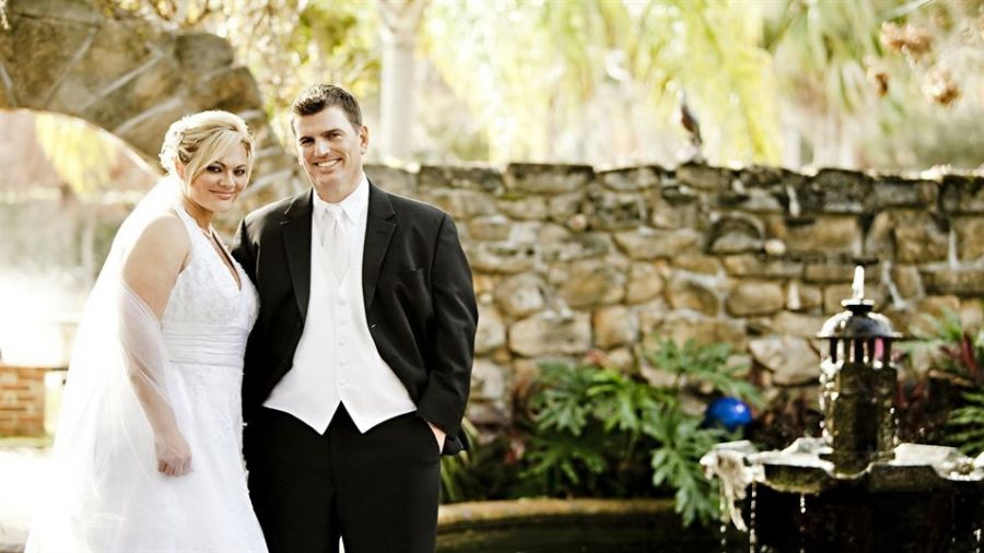 A joyous couple stands elegantly outside against a rustic stone wall and lovely fountain. The bride is dressed in a stunning white gown with a delicate veil while the groom looks sharp in his black suit and white vest. Sunlight filters through surrounding greenery, creating a beautiful outdoor wedding atmosphere filled with warmth and happiness.