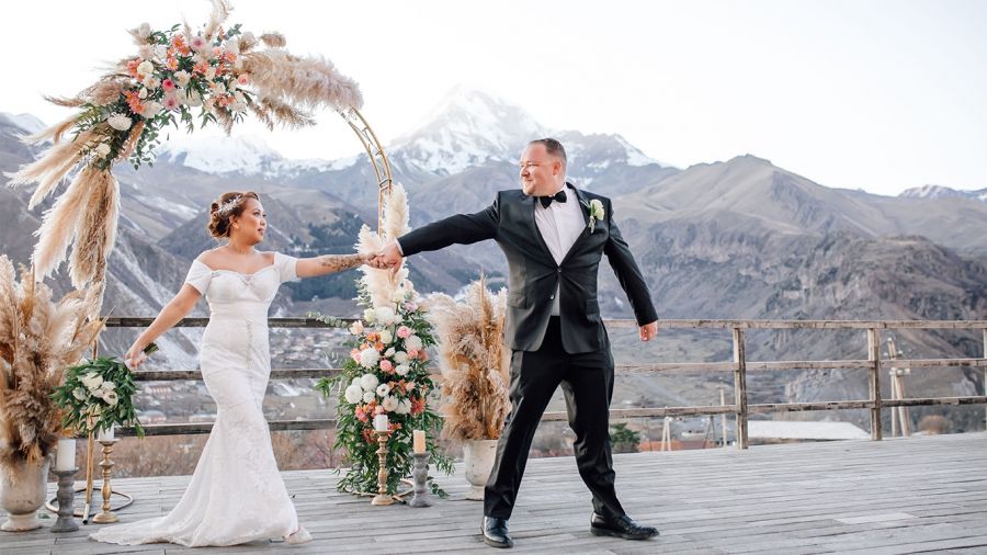 A happy couple stands outdoors on a wooden platform for their wedding ceremony. The bride wears an elegant white lace gown with a floral headpiece while the groom is dressed in a classic black tuxedo. Behind them, there is an arch of flowers and pampas grass framing the majestic snow-covered mountains under a bright blue sky creating a romantic atmosphere for their special day.