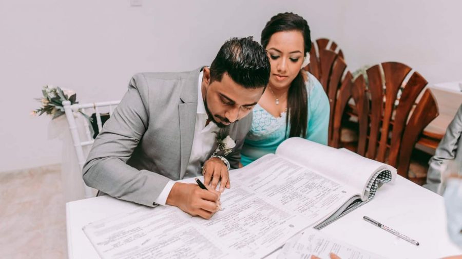 An image of a couple at their wedding ceremony shows them preparing to sign important documents. The man wears a gray suit while seated next to a woman in a light blue dress. They sit at a formal white table that holds an open ledger representing official documentation. The scene is set with minimal floral decor, creating an intimate atmosphere as they celebrate their love together through marriage.