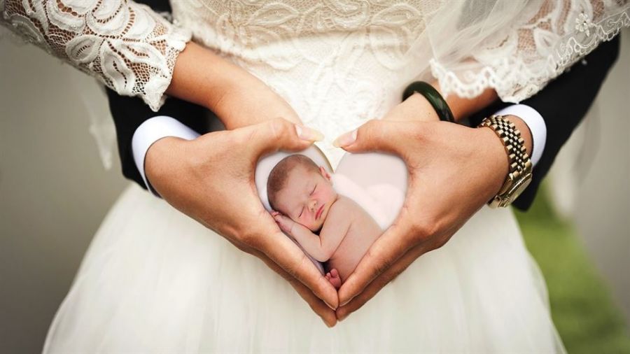 A couple dressed in wedding attire forms a heart shape with their hands, symbolizing love and unity. The woman wears a beautiful lace dress, while the man is in a sharp dark suit. The background features soft greenery that adds romance to the scene. Inside their heart-shaped hands is an edited image of a sleeping baby, representing family and new beginnings.