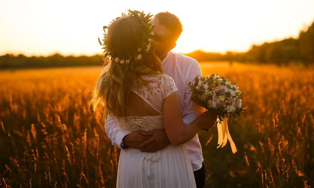 A couple embracing at sunset in a golden field, surrounded by tall grasses. The woman wears a delicate white dress and floral crown, holding a bouquet of flowers while the man dons a casual white shirt. The warm glow from the sun illuminates their joyful expression, creating an intimate atmosphere amidst soft silhouettes of trees against colorful skies.