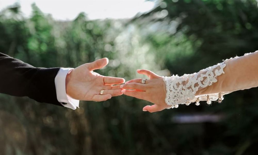 A close-up image of two hands reaching out towards each other against a blurred green background. One hand is dressed in a black suit while the other features an elegant lace sleeve. Both hands wear wedding rings, representing love and commitment.