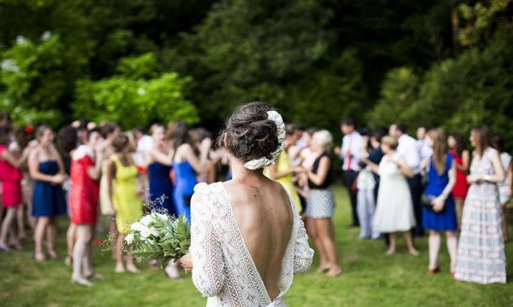 The image shows a bride in a lace dress holding a bouquet of white flowers facing away from the camera. Guests are mingling on lush green grass, enjoying the festivities of an outdoor wedding. The ambiance is vibrant with colorful attire worn by attendees who celebrate joyfully in a beautiful garden setting, embodying romance and happiness during this special occasion.