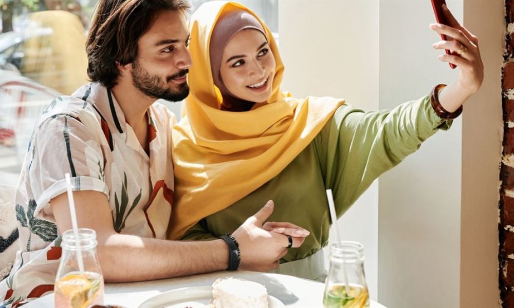 Legal Requirements Of A Wedding In Abu Dhabi Vs Dubai 2Fffbbb2 | Everything You Need To Know About: Legal Requirements Of A Wedding In Abu Dhabi Vs Dubai | Are You Confused About Your Wedding In Abu Dhabi; And Would Love To Get Some Assistance. Do Not Hesitate To Book A Free Consultation From Our Team Of Experts. A happy couple sits at a brightly lit table enjoying drinks together. The woman is wearing a colorful headscarf and green top while holding a phone as if taking a selfie. The man has medium-length hair and a patterned shirt. They look relaxed smiling at each other in a cozy dining spot with brick walls, creating an inviting atmosphere perfect for sharing joyous moments.