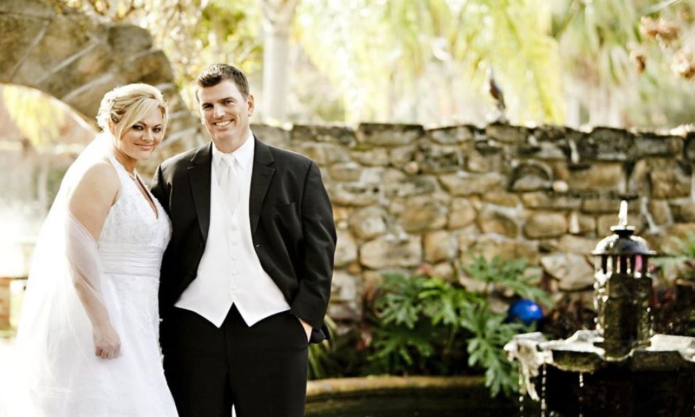 A joyous couple stands elegantly outside against a rustic stone wall and lovely fountain. The bride is dressed in a stunning white gown with a delicate veil while the groom looks sharp in his black suit and white vest. Sunlight filters through surrounding greenery, creating a beautiful outdoor wedding atmosphere filled with warmth and happiness.
