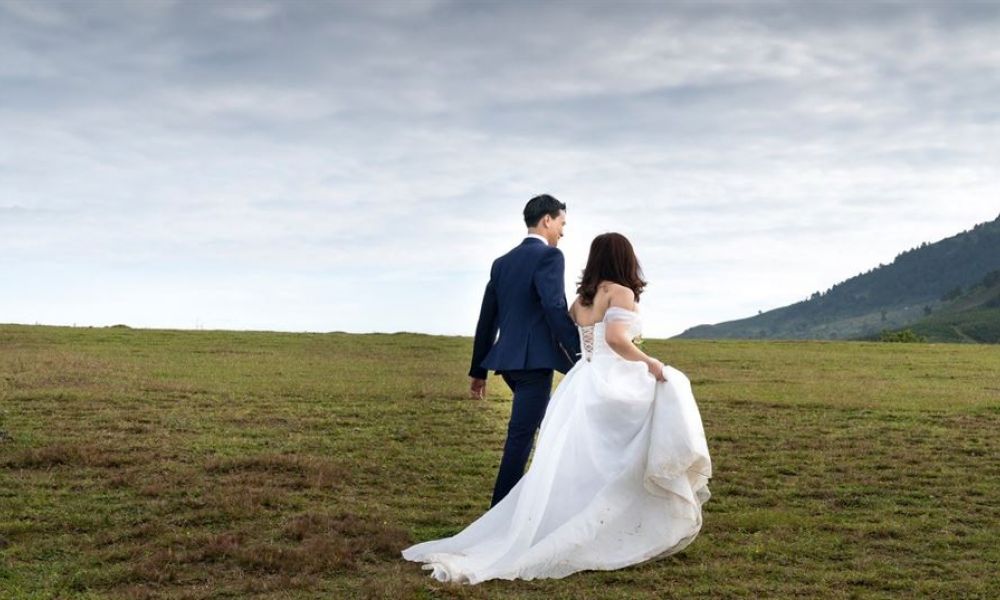 The image captures a couple joyfully walking hand in hand on a grassy landscape. The bride is adorned in a white gown with a long flowing train while the groom wears a dark blue suit. Their surroundings feature green rolling hills under an idyllic cloudy sky, creating a romantic outdoor atmosphere that symbolizes everlasting love.