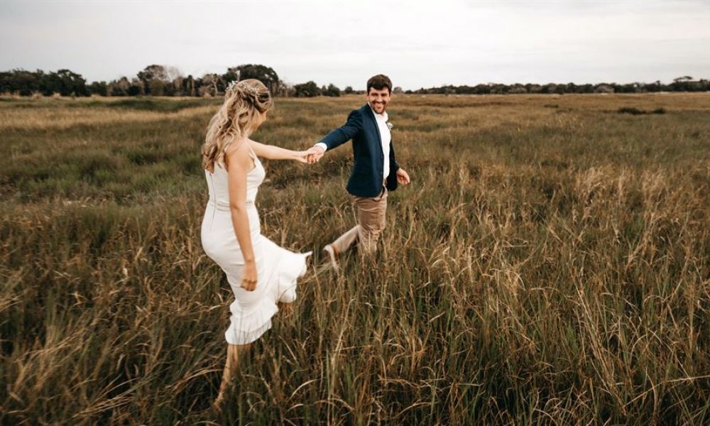 Why Dubai Residents Must Consider An Easy Wedding In Georgia 54732402 | Why Dubai Residents Must Consider An Easy Wedding In Georgia | Why Is It A Good Idea To Hold A Wedding In Georgia, You Might Wonder? The image showcases a happy couple enjoying nature together in wide open grasslands under a cloudy sky. The woman wears a beautiful white dress while the man is dressed smartly in blue and light pants. They are holding hands, reflecting love and joy as they share this moment outdoors. The serene setting adds charm to their adventure-like appearance amidst tall grass and gentle vegetation.