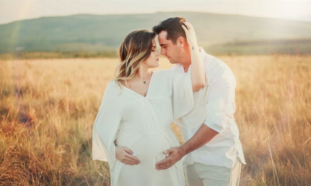 A couple stands lovingly in a golden field during sunset, holding each other with joy glowing on their faces. The woman wears a flowing white dress while she cradles her pregnancy bump, and the man gently rests his hand on it. They gaze into each other’s eyes, capturing a moment filled with love amidst a serene landscape of hills and blue skies. The intimate scene reflects warmth, connection, and anticipation for their shared future together.