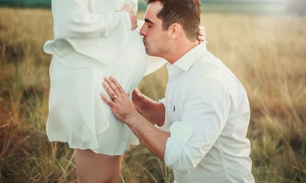An image of a couple outdoors shows a man tenderly kissing the belly of a pregnant woman. They wear elegant white outfits amidst tall grass in soft sunlight, radiating love and serenity.