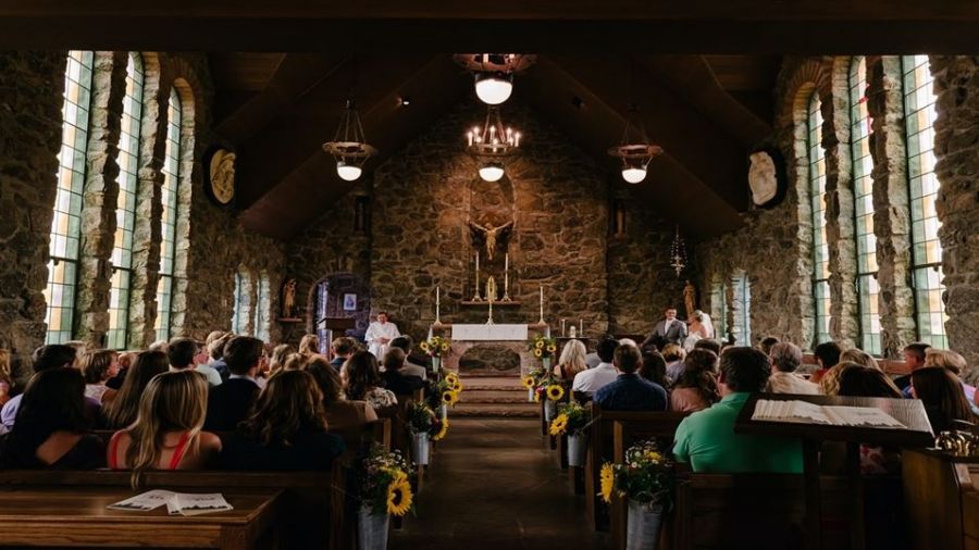 The image shows a lovely church filled with guests enjoying a wedding ceremony. Sunflowers line the aisle while light streams through arched windows enhancing the joyous atmosphere. An officiant stands at an altar adorned with candles symbolizing love and unity making this a heartwarming moment for everyone involved.