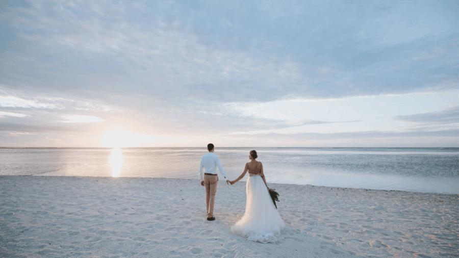 A couple walks hand in hand on a serene beach during sunset. The bride’s flowing white gown contrasts beautifully with the groom's light-colored outfit. Soft clouds float across the sky as they approach the warm ocean water, creating an ambiance of romance and tranquility perfect for weddings or intimate moments. The wide sandy beach surrounds them while the colors of sunset illuminate this peaceful scene inviting couples to create their own wedding dream here in Dubai.
